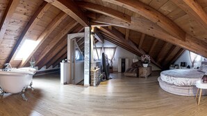 Een kamer onder het dak met houten balken, een badkuip, een bed en een bureau.