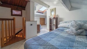 Una camera da letto con una scala in legno, un letto con una coperta blu, un piccolo armadietto bianco e un lucernario.