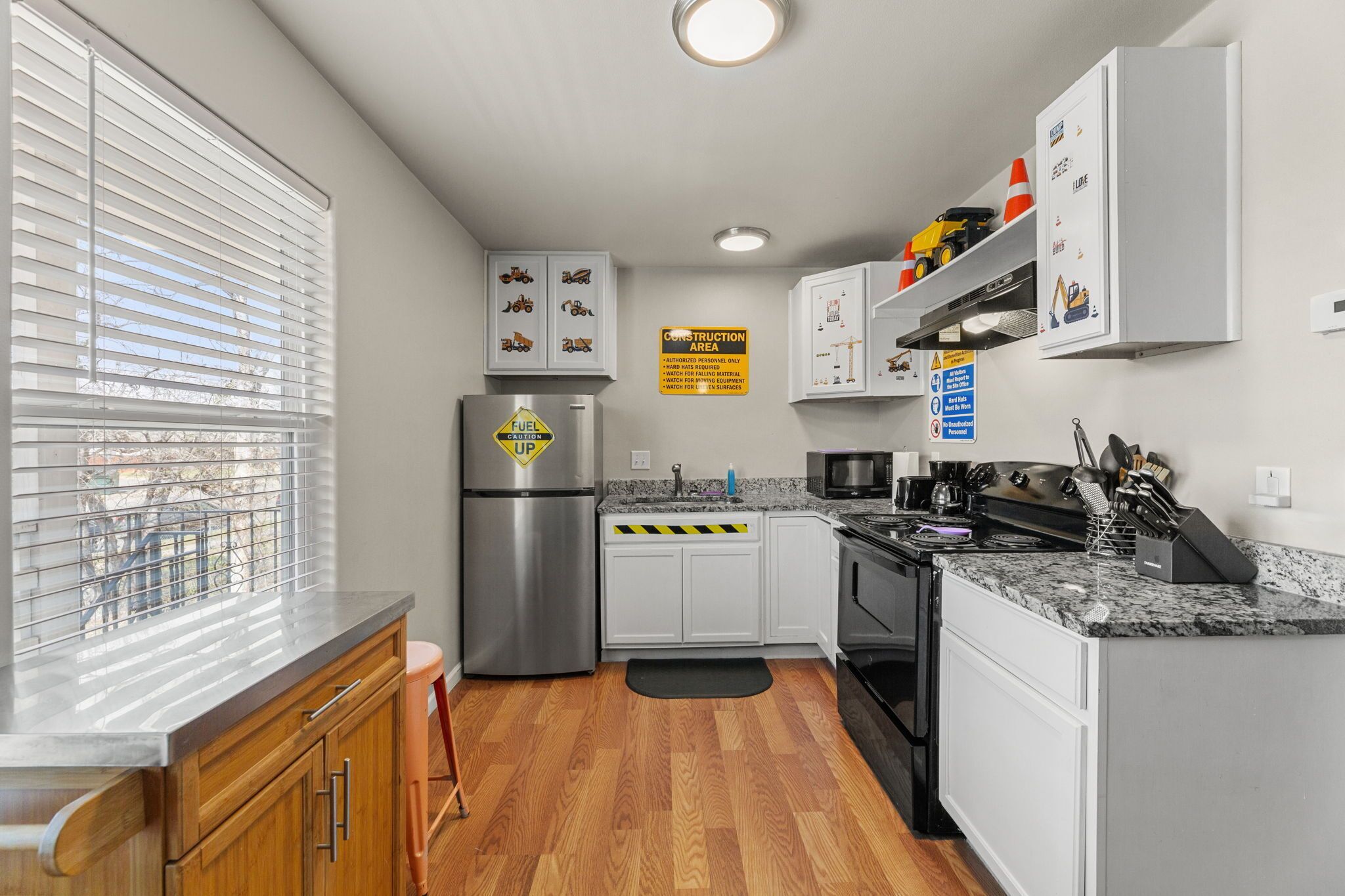 A modern kitchen with white cabinets, a stainless steel refrigerator, granite countertops, and a window with blinds.