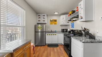 A modern kitchen with white cabinets, a stainless steel refrigerator, granite countertops, and a window with blinds.