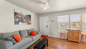 Ein Wohnzimmer mit grauem Sofa, orangefarbenen Kissen, einem schwarzen Couchtisch, einem Deckenventilator und einem großen Fenster mit Jalousien.