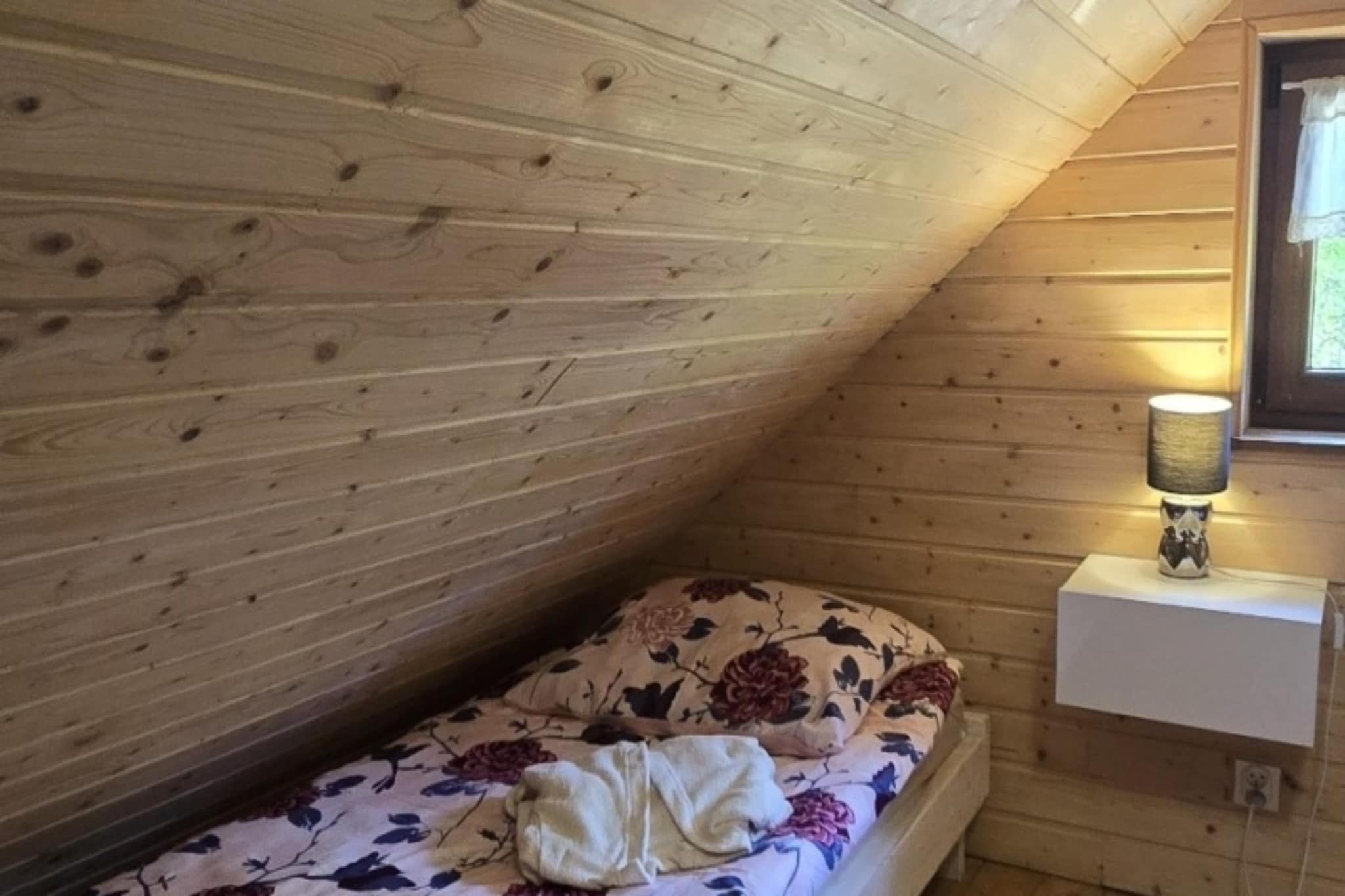 Ein kleiner Raum mit schrägem Dach, Holzwänden, einem Einzelbett mit floralem Bettbezug und einer Nachttischlampe.