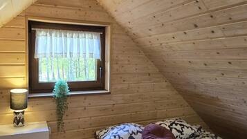 Un dormitorio acogedor en el ático, con techo inclinado, ventana con cortina, lámpara de noche y colcha estampada.