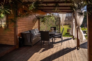 A covered outdoor seating area with a wooden deck, a wicker sofa, a dining table, and chairs.