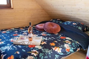 A bed with a floral bedspread, two pillows, a tray with a bottle and glasses, and a window in a wooden room.