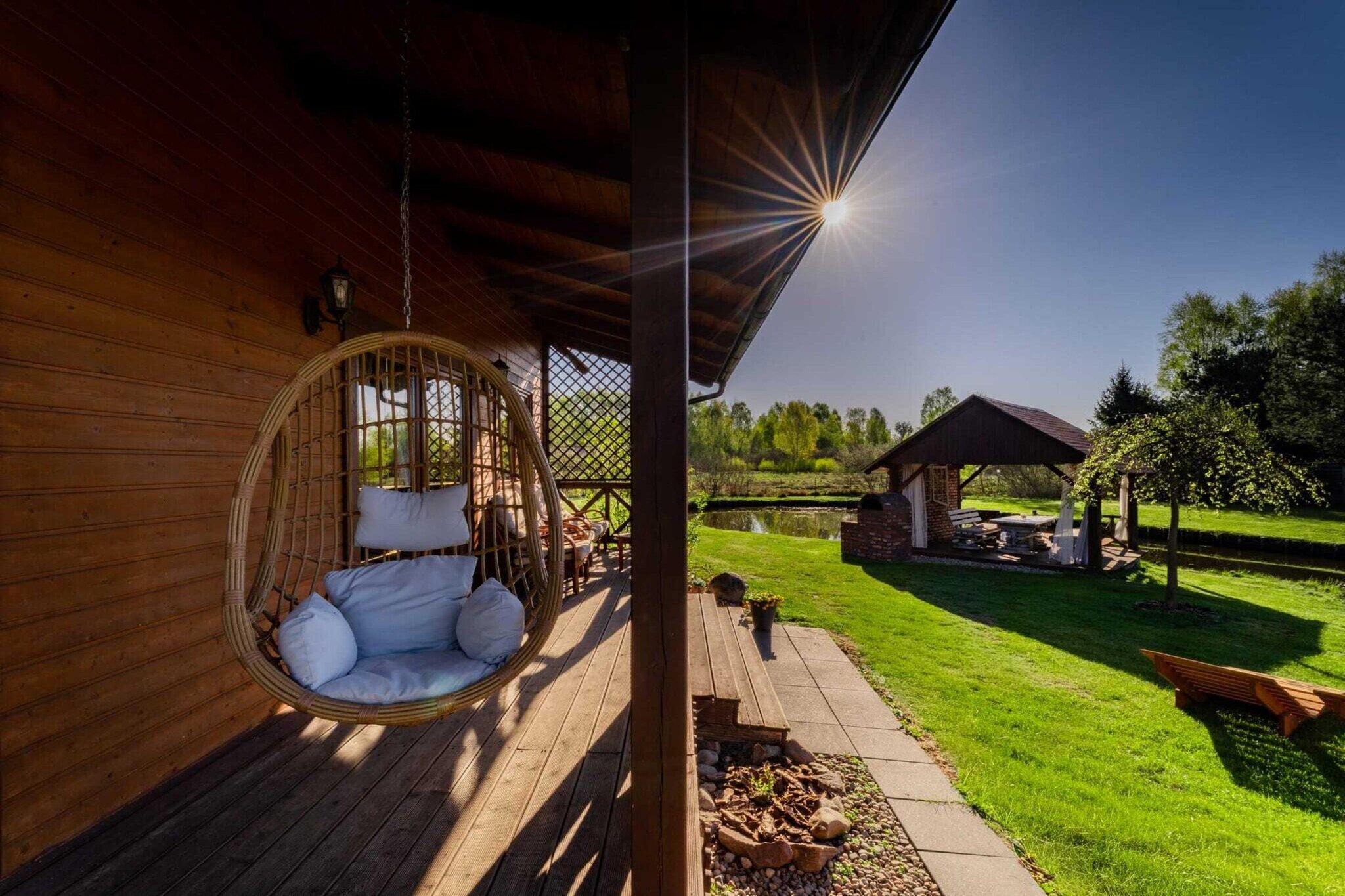 Una terraza de madera con una hamaca colgante, zona de barbacoa y vistas a un estanque y árboles.