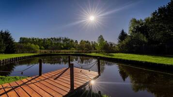 En träbrygga som sträcker sig ut över en stilla vattenyta, omgiven av grönska och träd under en klar himmel.