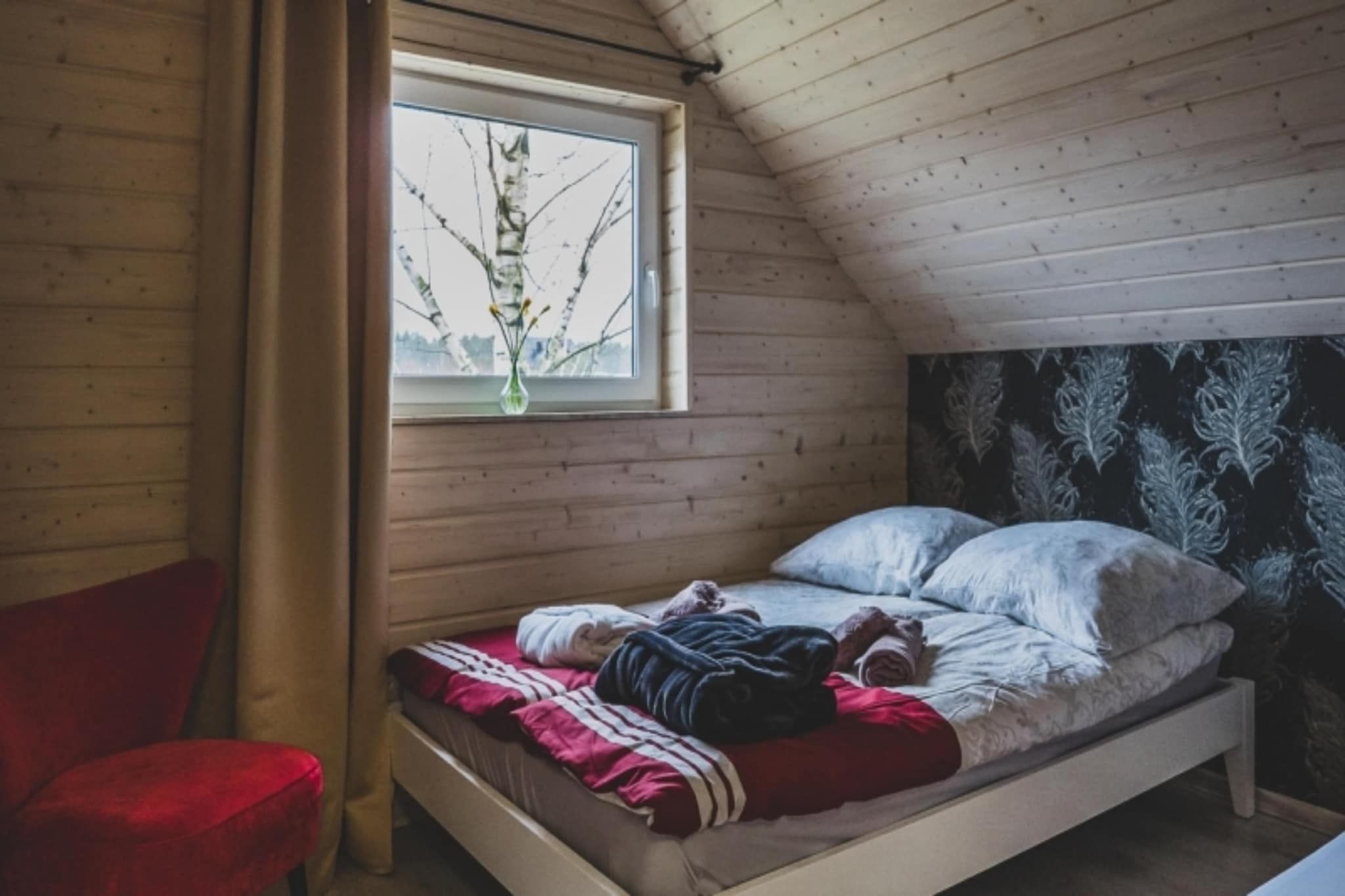 A single bed with pillows and a blanket, a window with a plant, and a red chair in a wooden room.