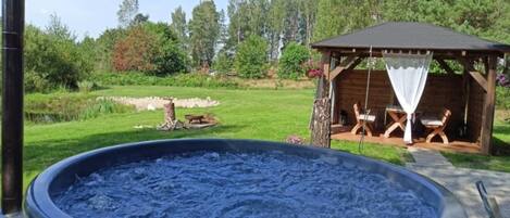 An outdoor hot tub with a wooden deck, surrounded by a grassy area and a wooden gazebo with seating.