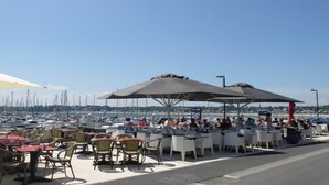 Ein Essbereich im Freien mit Tischen, Stühlen und Sonnenschirmen, mit Blick auf den Hafen im Hintergrund.