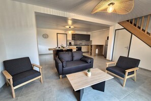 A modern living room with a sofa, two armchairs, a coffee table, and a kitchen in the background. - Terraced Country House near Sea (Plurien)