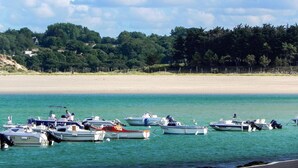 Ein Strand mit vor Anker liegenden Booten in ruhigem Wasser, einem sandigen Ufer und einer bewaldeten Hügelkette im Hintergrund.
