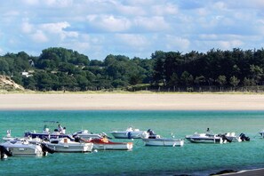 A beach with boats docked in calm waters, a sandy shore, and a forested hill in the background. - Terraced Country House near Sea (Plurien)