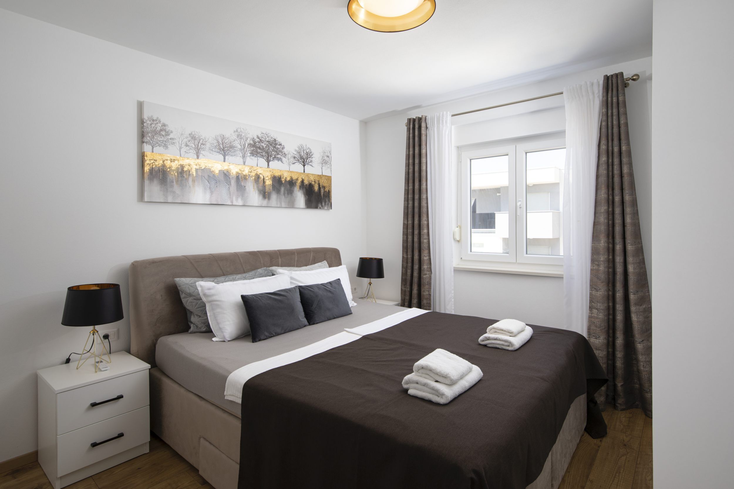Een slaapkamer met een bed, nachtkastjes, een lamp, een schilderij, een raam met gordijnen en een uitzicht op gebouwen.