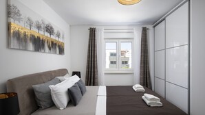 A modern hotel room with a bed, a painting, a window with curtains, and a view of buildings outside. - Luxury Penthouse Pula (Pula)