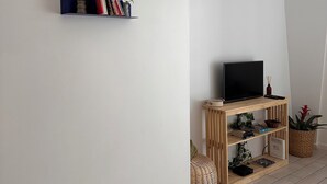 Een woonkamer met een flatscreen televisie op een houten standaard, een boekenkast met boeken en een klein plantje, en een rieten mand op de vloer.