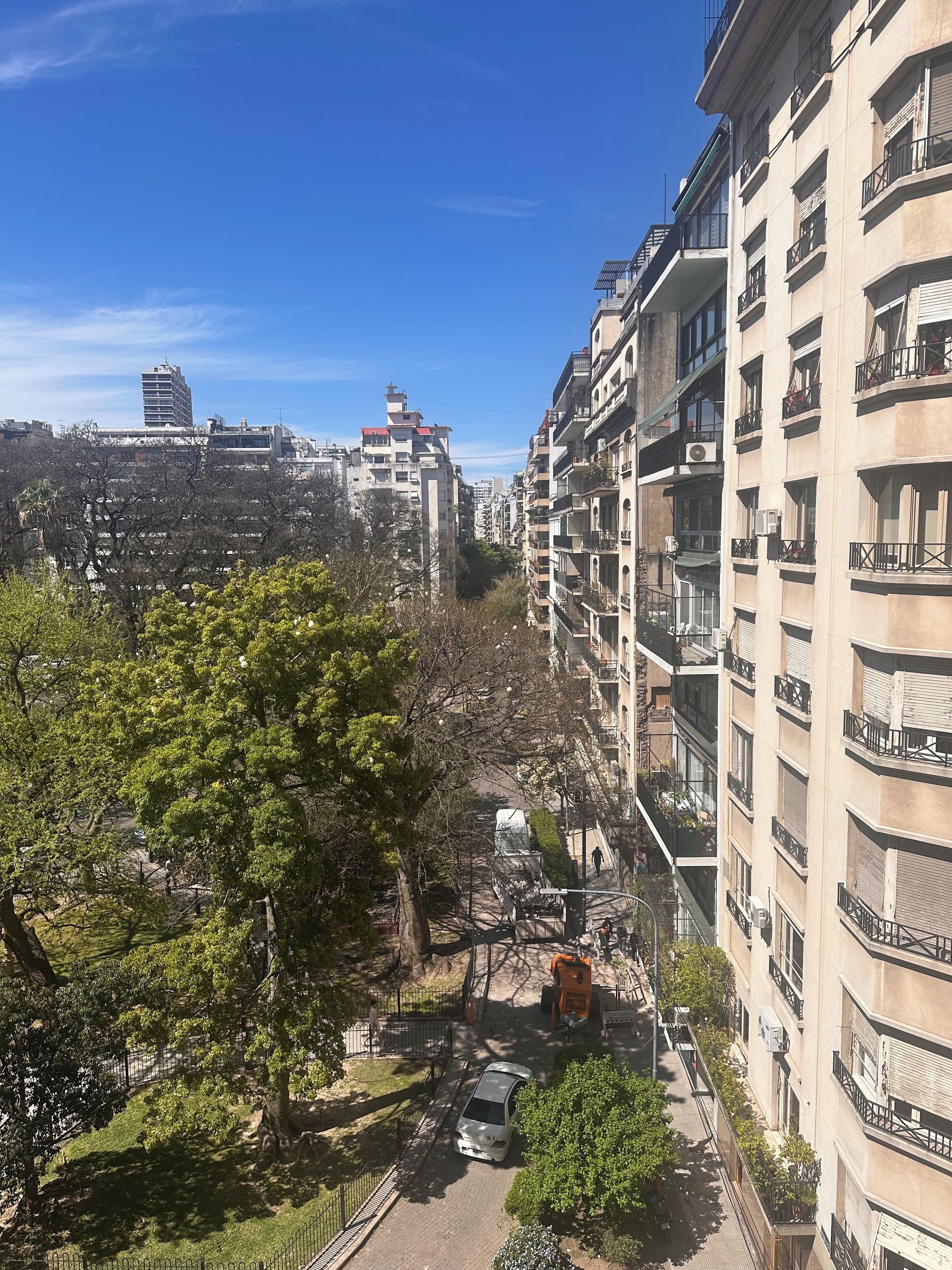 Un edificio residencial de varios pisos con balcones, cielo despejado y una calle bordeada de árboles.