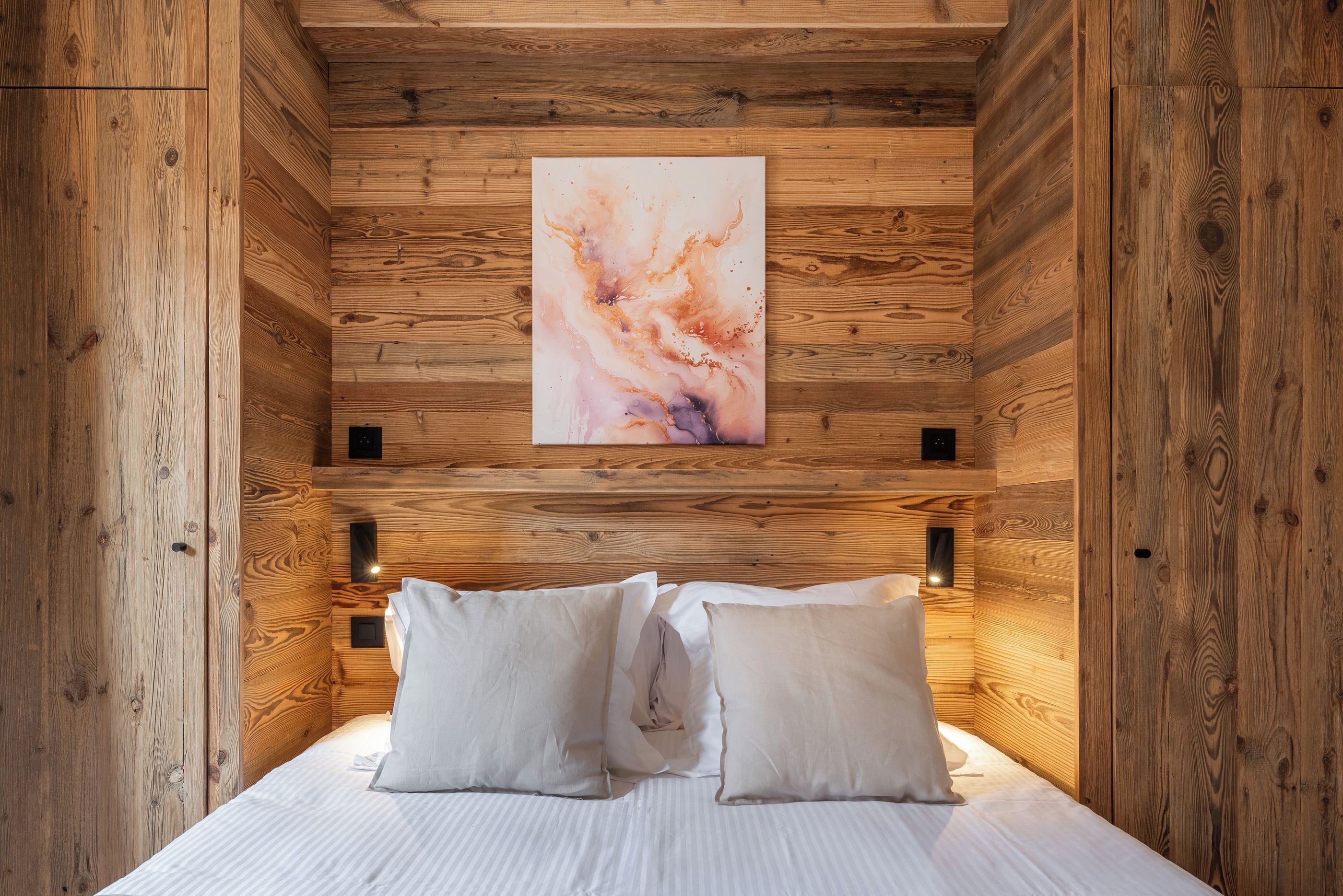 A wooden cabin room with a bed, pillows, a wall-mounted abstract painting, and built-in lighting.