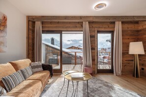 Een gezellige woonkamer met een houten plafond, een bank met gestreepte kussens, een klein rond tafeltje en uitzicht op een besneeuwd berglandschap door grote schuifdeuren.