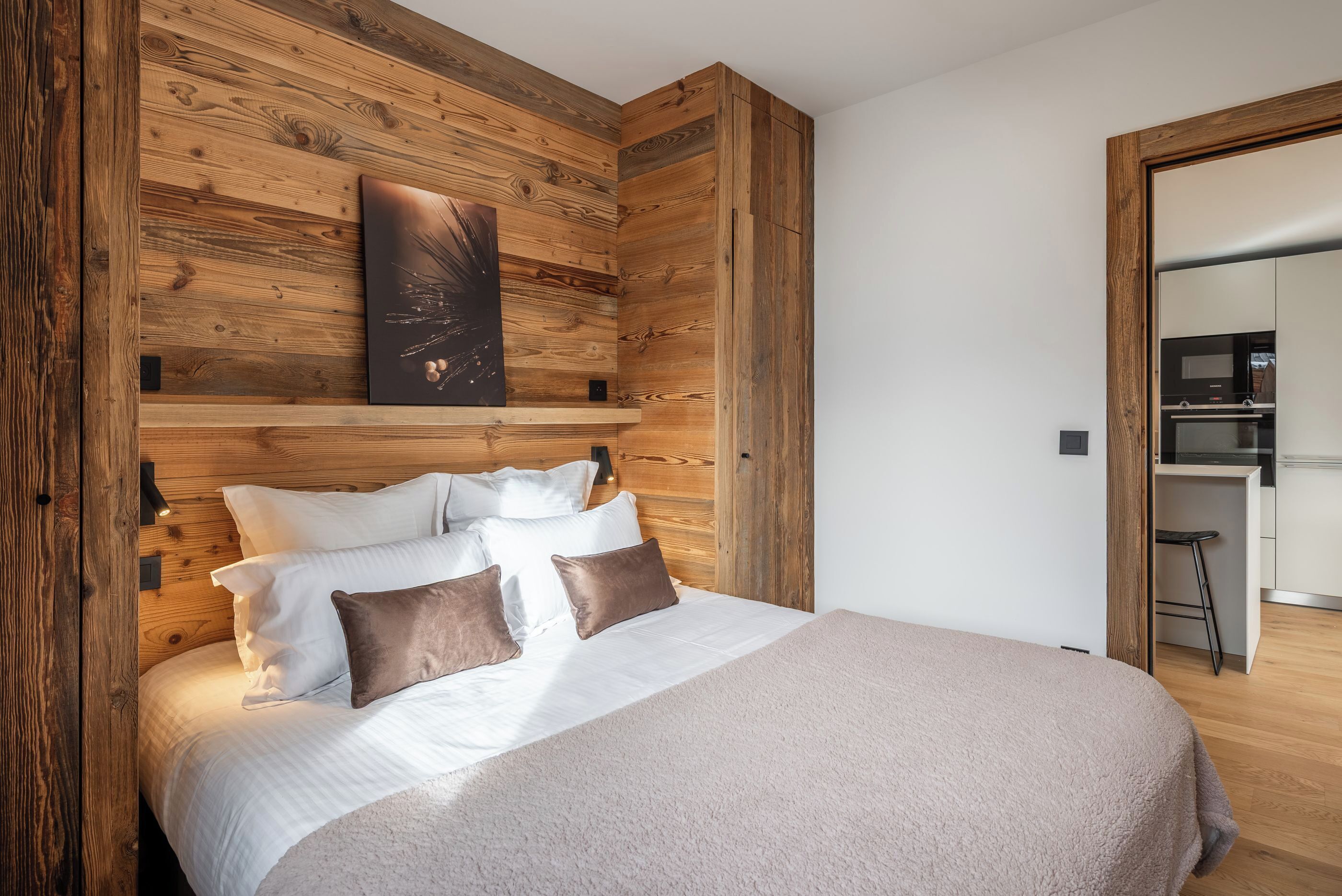 Un dormitorio con cabecera de madera, cama con ropa de cama blanca y marrón, un cuadro en la pared y una cocina visible a través de una puerta abierta.