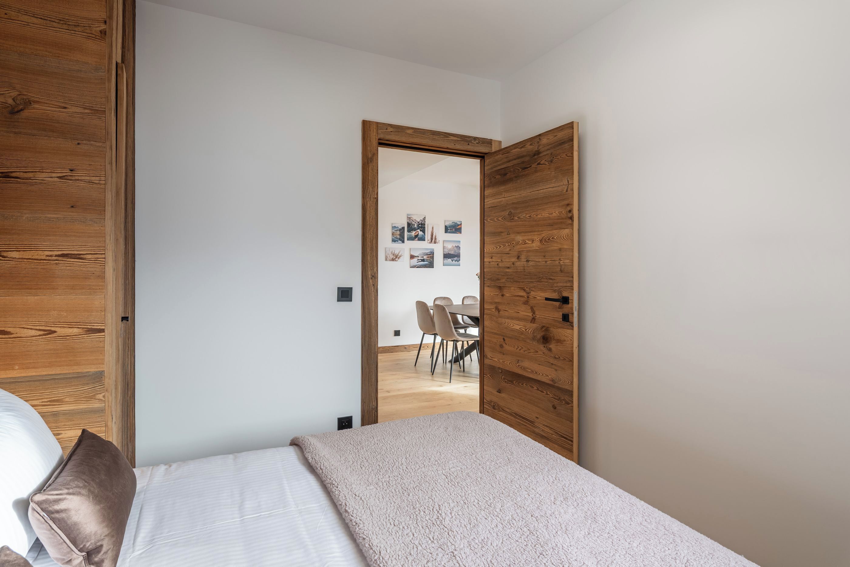 A bedroom with a bed, wooden wardrobe, and a door leading to a dining area with chairs and a table.