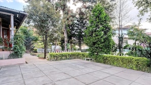 Um pátio pavimentado com piso de azulejos, rodeado por vegetação e um edifício com entrada coberta.