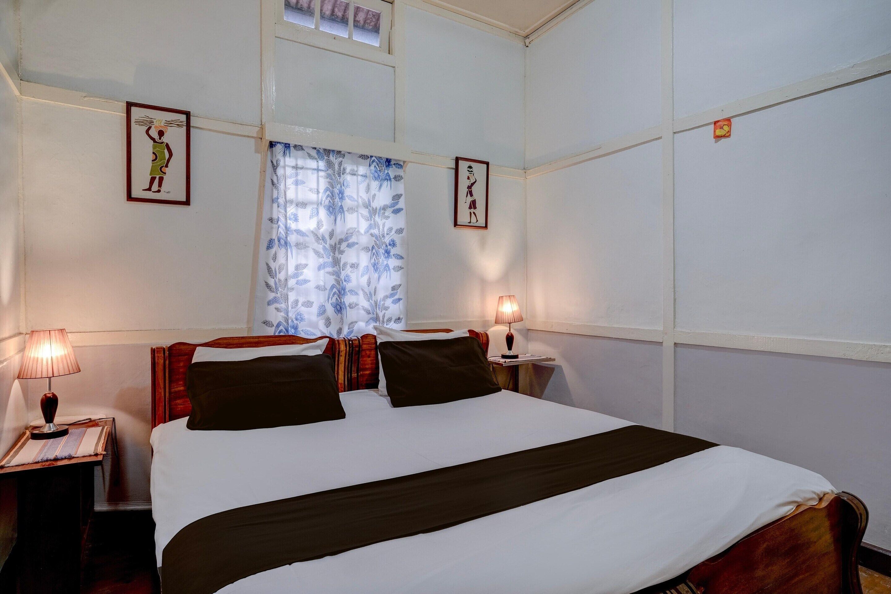 A neatly made bed with a white and brown bedspread, two bedside lamps, a window with blue curtains, and two framed pictures on the wall.