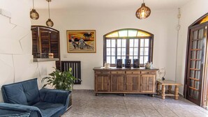 Uma sala de estar com um sofá azul, um armário de madeira com caixas de som, um quadro e uma janela com vista.