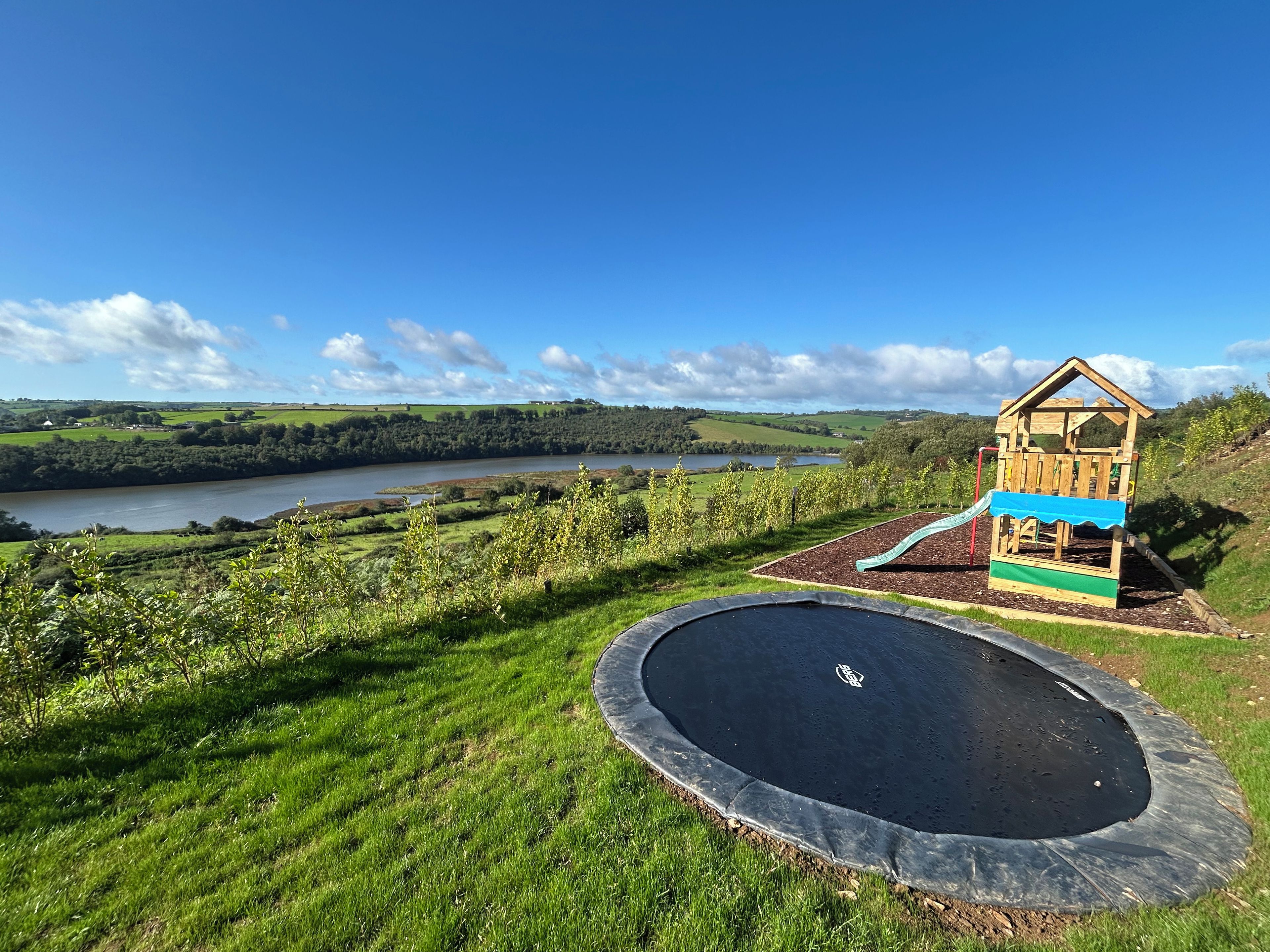 A playground with a slide and a wooden playhouse, surrounded by a grassy area and a body of water in the background.