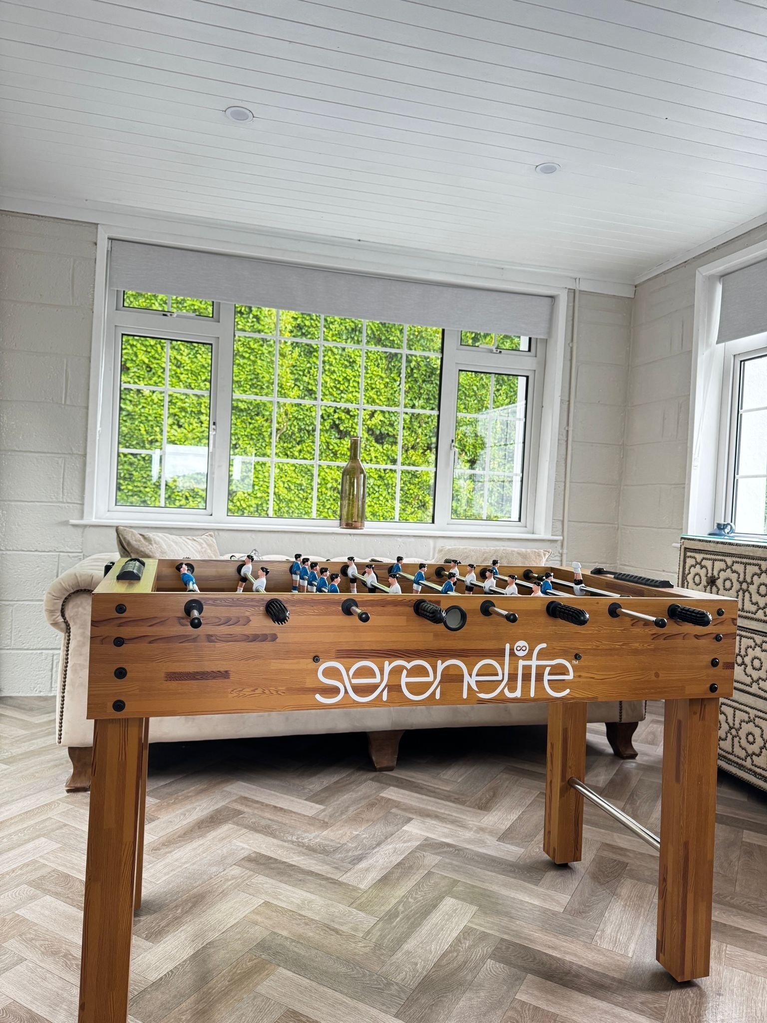 A foosball table with the brand 'serenelife' in a room with a herringbone floor and a window with a view of greenery.