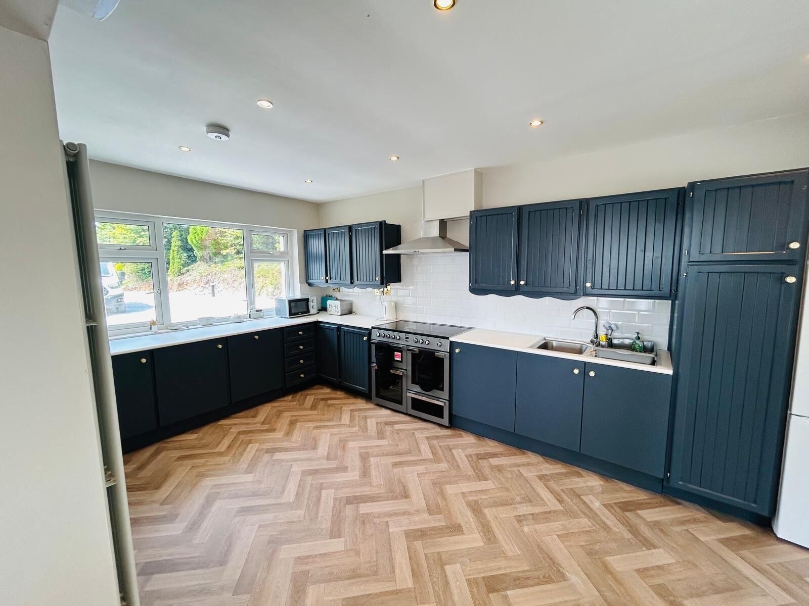 A modern kitchen with dark blue cabinets, a herringbone wood floor, and a window overlooking greenery.