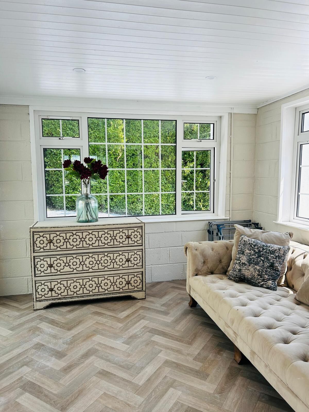 Une pièce dotée d’un parquet à motifs en chevrons, d’un canapé beige agrémenté d’un coussin à motifs, d’une commode décorative et d’une grande fenêtre donnant sur un paysage verdoyant.