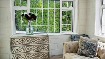 Une pièce dotée d’un parquet à motifs en chevrons, d’un canapé beige agrémenté d’un coussin à motifs, d’une commode décorative et d’une grande fenêtre donnant sur un paysage verdoyant.