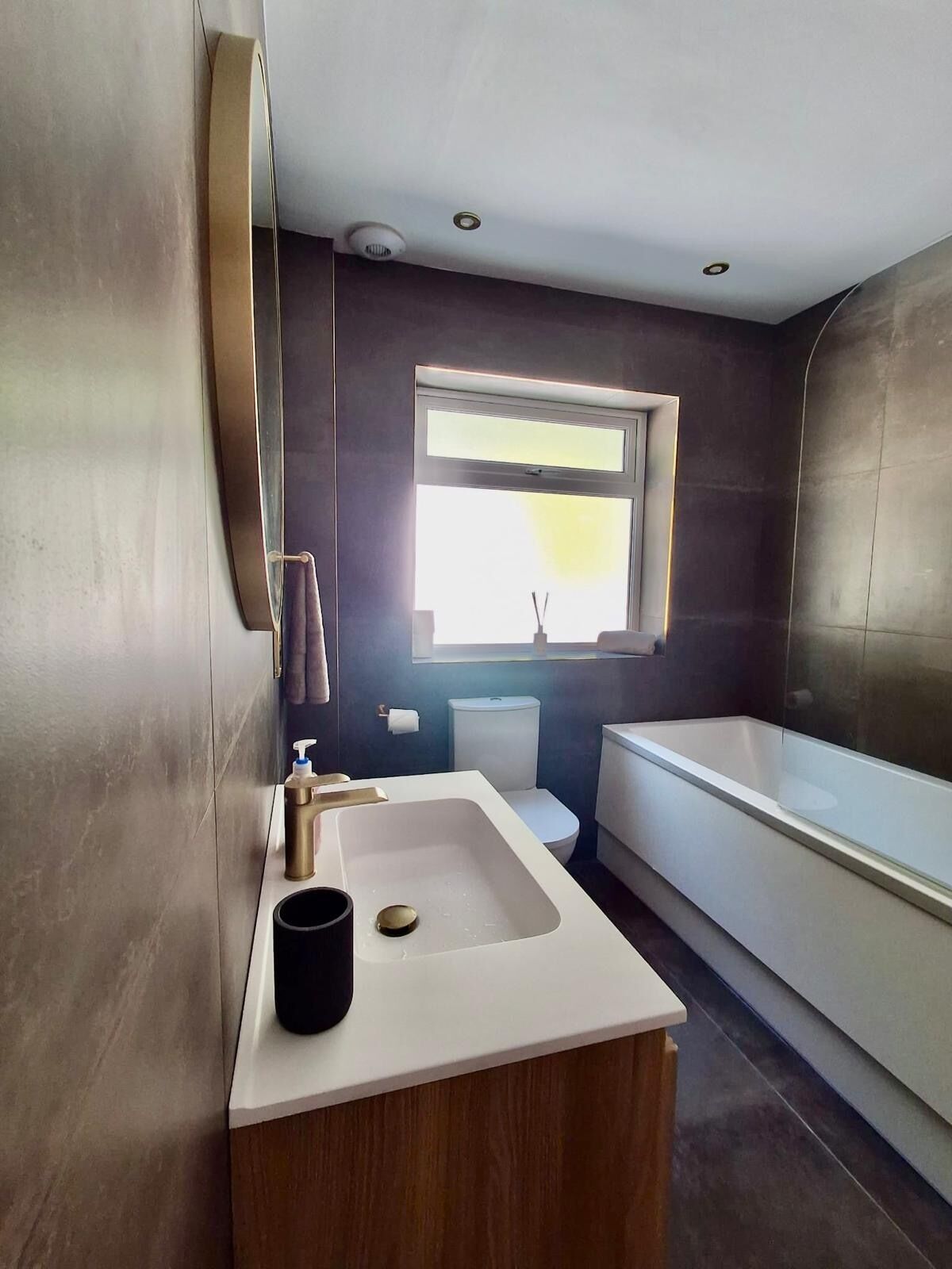 A modern bathroom with a large bathtub, a sink with a wooden vanity, a toilet, and a window above the sink.