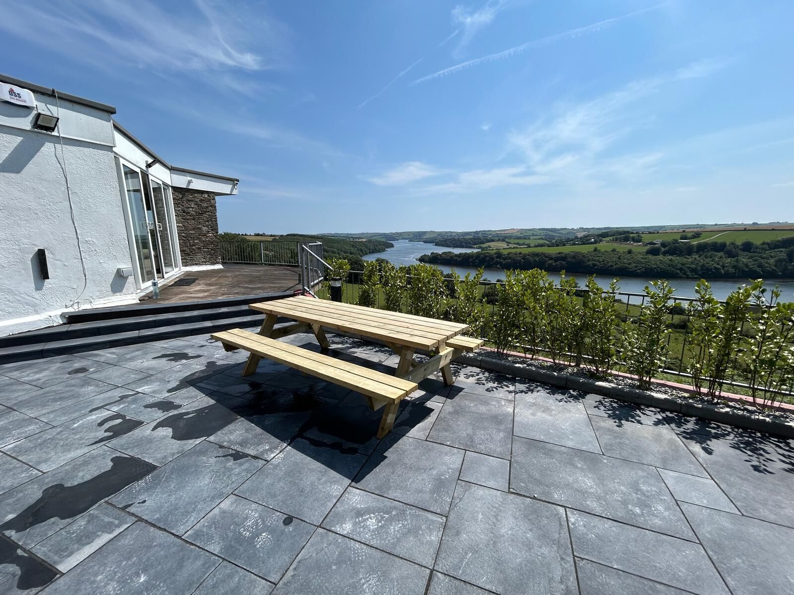 A patio with a wooden table and benches overlooking a river and green landscape.