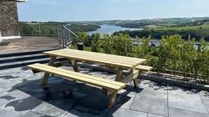 Une table de pique-nique en bois, posée sur une terrasse en pierre, d’où l’on a vue sur un lac et des collines.