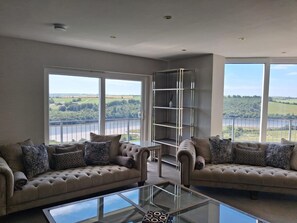 A spacious living room with two sofas, a glass coffee table, and a metal shelving unit. Large windows offer a view of a river and greenery. - The Waterfront House 9 minutes drive to Kinsale,6 bedrooms , sleeps 19 guests. (Cork)