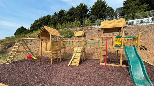 A playground with wooden structures, including a slide and climbing areas, set on mulch ground.