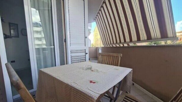 A small balcony with a table and chairs, a sliding glass door, and a striped awning. - Lights on the Sea (Alghero)