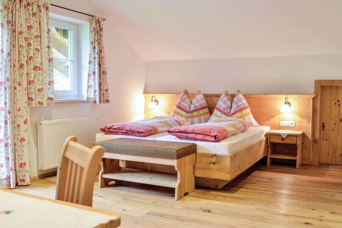 Ein gemütliches Schlafzimmer mit einem hölzernen Bett, einem Nachttisch, einem Sessel und einem Fenster mit blumigen Vorhängen.