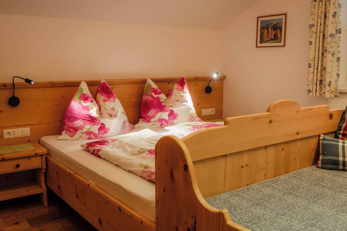 Ein hölzernes Bett mit floraler Bettwäsche, ein Nachttisch, eine Lampe und ein Bild an der Wand.