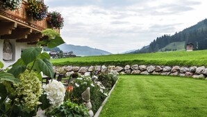 Een hotel met een stenen muur, een houten balkon met bloembakken en uitzicht op een groen veld en bergen.