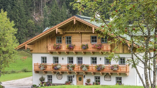Een houten chalet met twee verdiepingen en balkons versierd met bloembakken, gelegen tegen een achtergrond van bos.