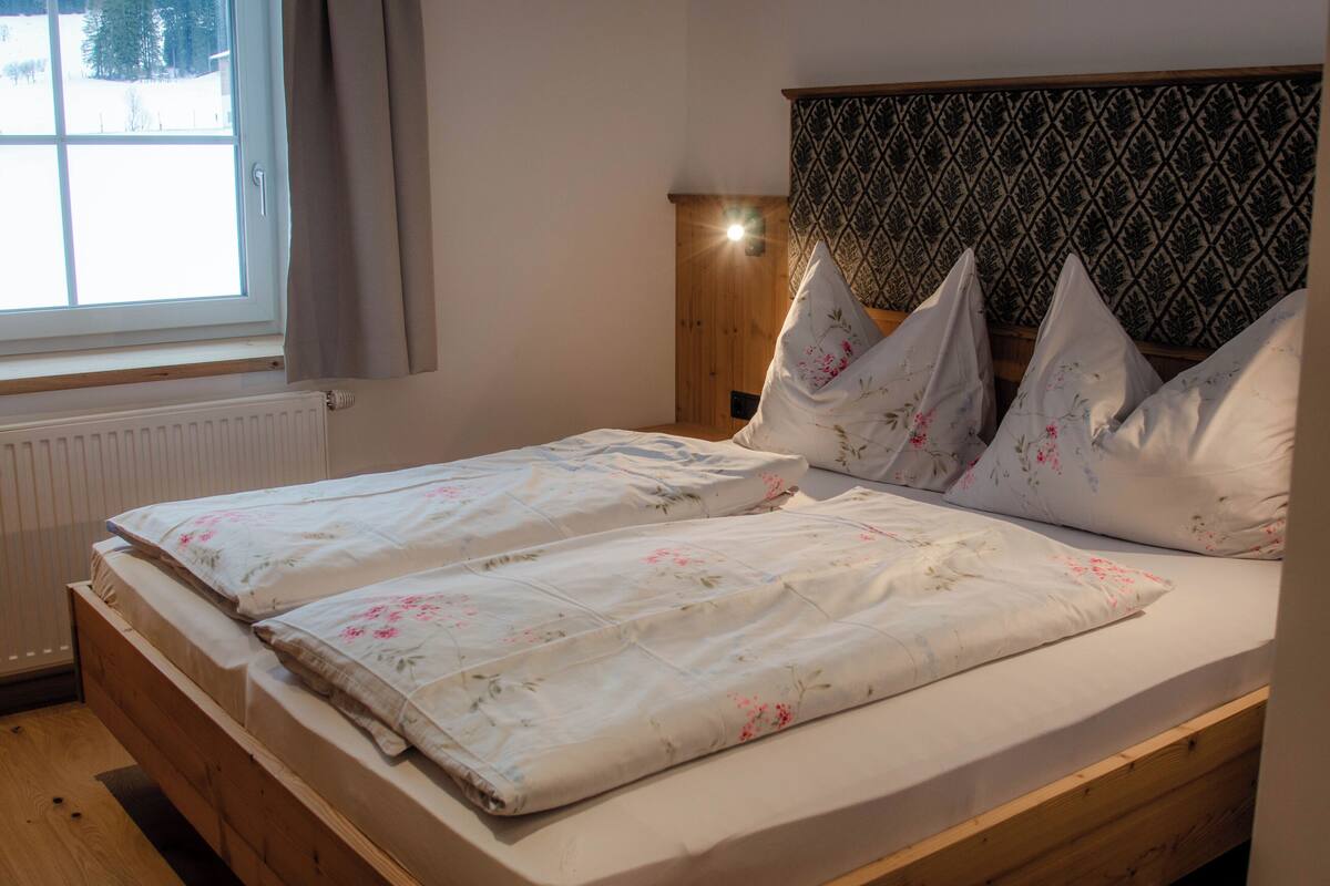 Ein Doppelbett mit floralem Bettbezug, ein Holz-Kopfteil und ein Fenster mit Vorhängen.