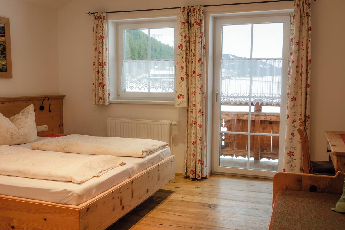 Ein Schlafzimmer mit zwei Betten, einem Holz-Kopfteil, einem Fenster mit floralem Vorhang und Blick auf eine Holzterrasse.