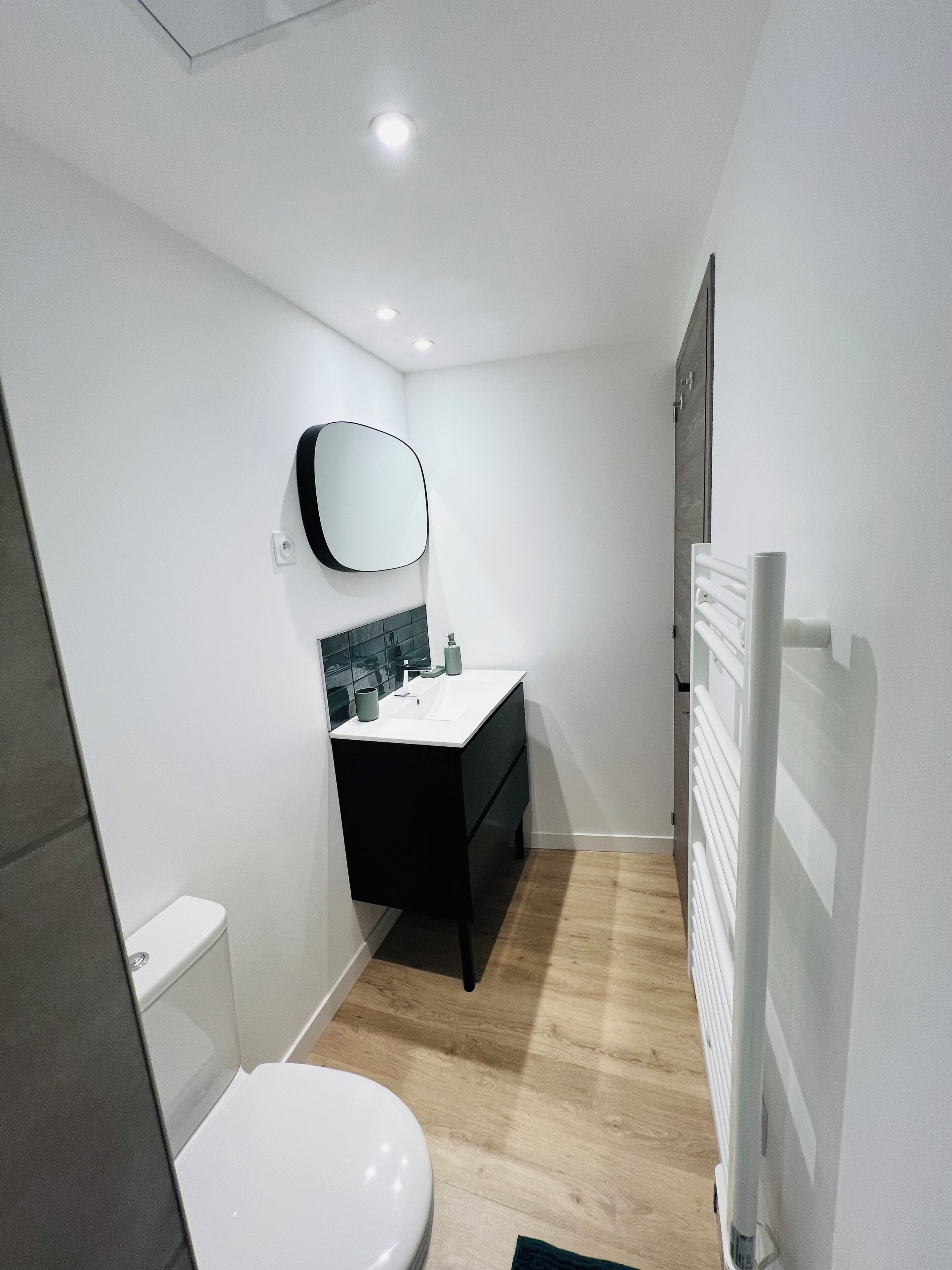Une salle de bain moderne avec des toilettes blanches, un meuble-lavabo noir doté d’un miroir et un radiateur blanc.