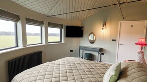 A bedroom with a large bed, a desk, a mirror, a TV, and a window with a view of a field. - The Corn Silo (Ipswich)
