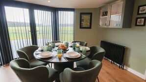 A dining area with a round table set for six, surrounded by green upholstered chairs. A breakfast setting includes plates, cups, and a croissant. The room features a large window with blinds, a radiator, and a framed quote on the wall. - The Corn Silo (Ipswich)