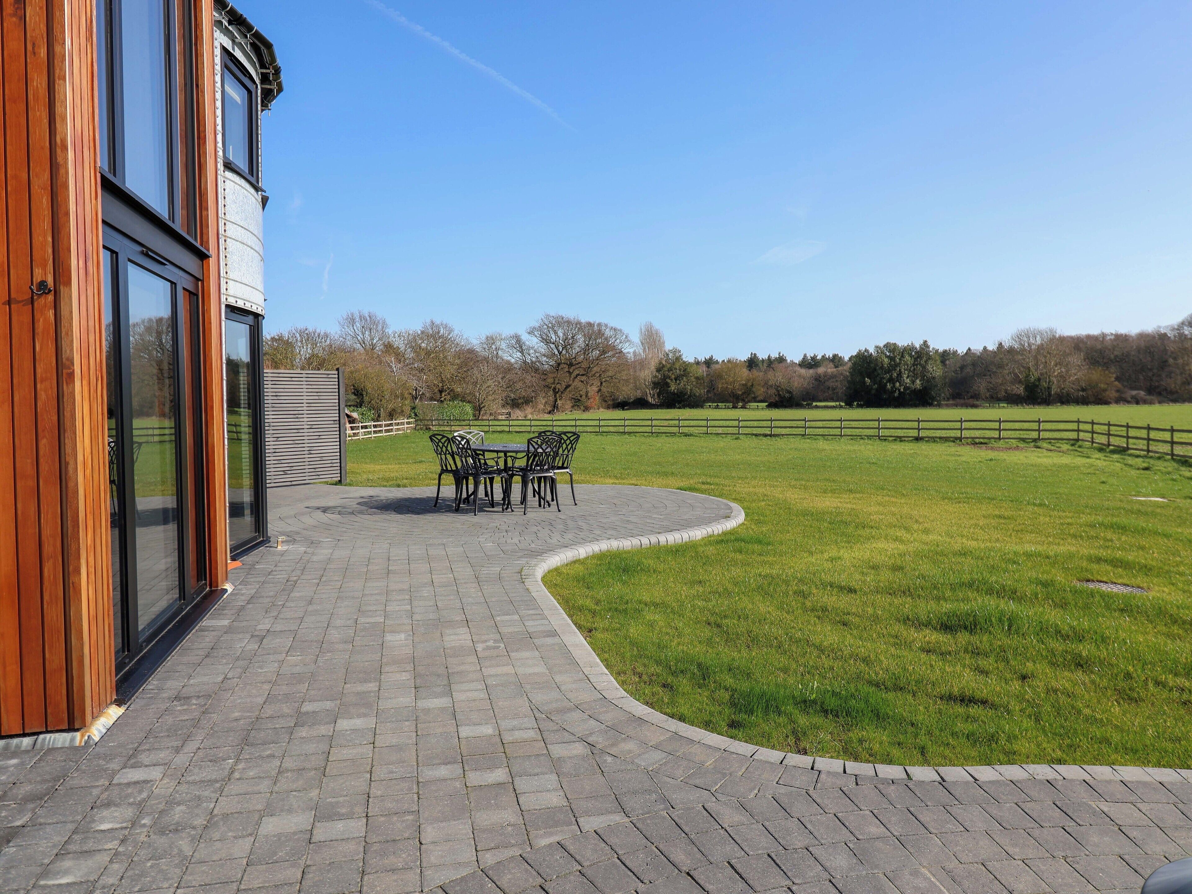 A patio with a brick pathway leading to a table and chairs, surrounded by a grassy field and a wooden fence.