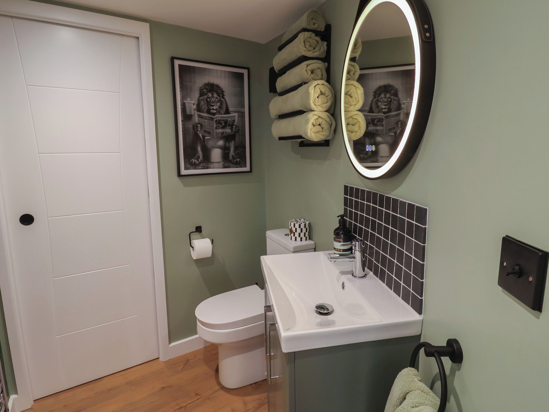 Une salle de bain avec des toilettes, un lavabo et un miroir avec un éclairage intégré. On y trouve des serviettes sur un porte-serviettes et un tableau encadré accroché au mur.